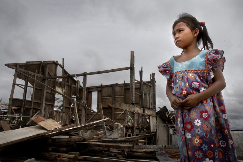A little girl, Eden Fe, stands outside a ruined house.