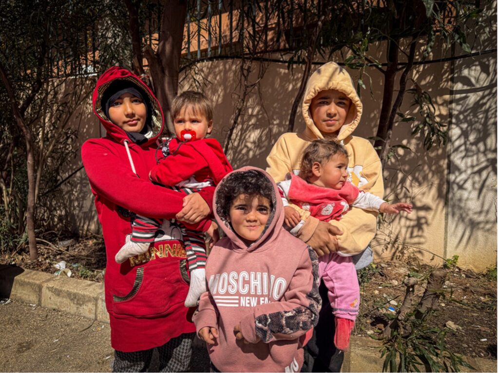 Four children stand in the shade against a wall. Two of the older children hold an infant each